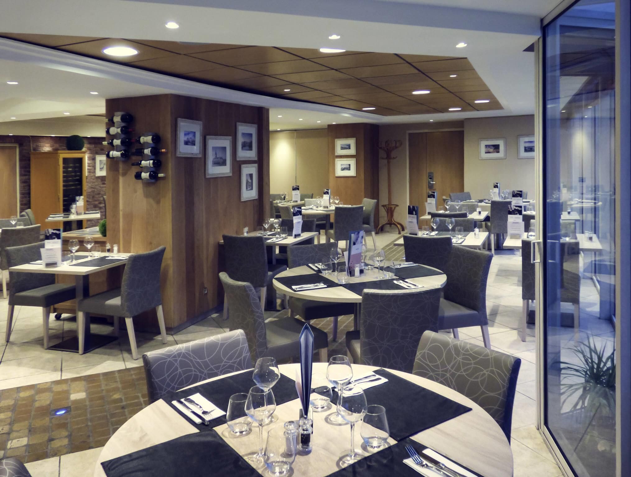 A modern restaurant with round tables, grey upholstered chairs, and a wooden accent wall.