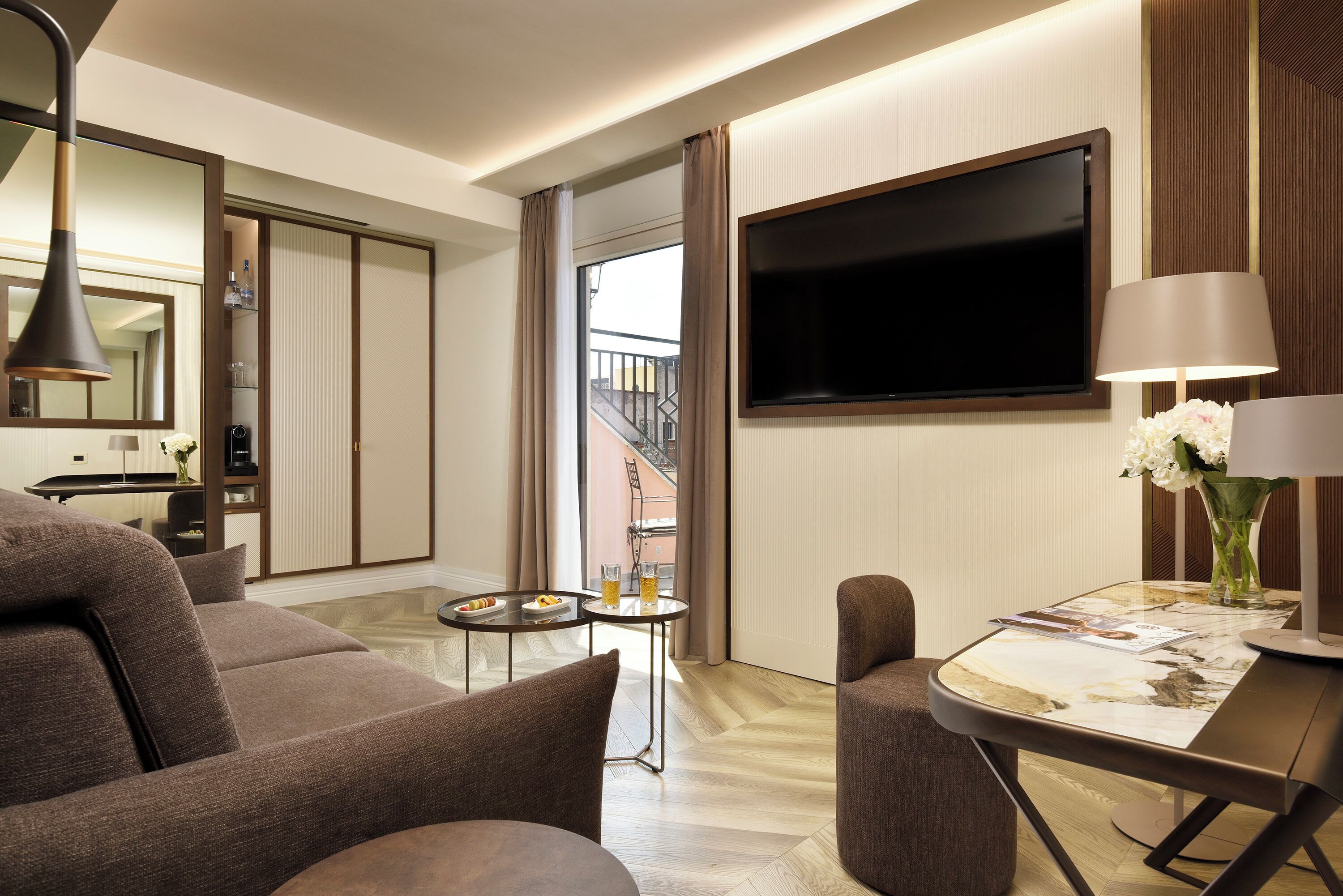 A modern hotel room with a sofa, a small dining table, a TV mounted on the wall, and a balcony visible through the door.