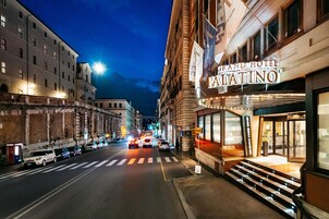A street view at night with a Grand Hotel Piatino on the right, featuring a prominent entrance and a staircase leading to the lobby.