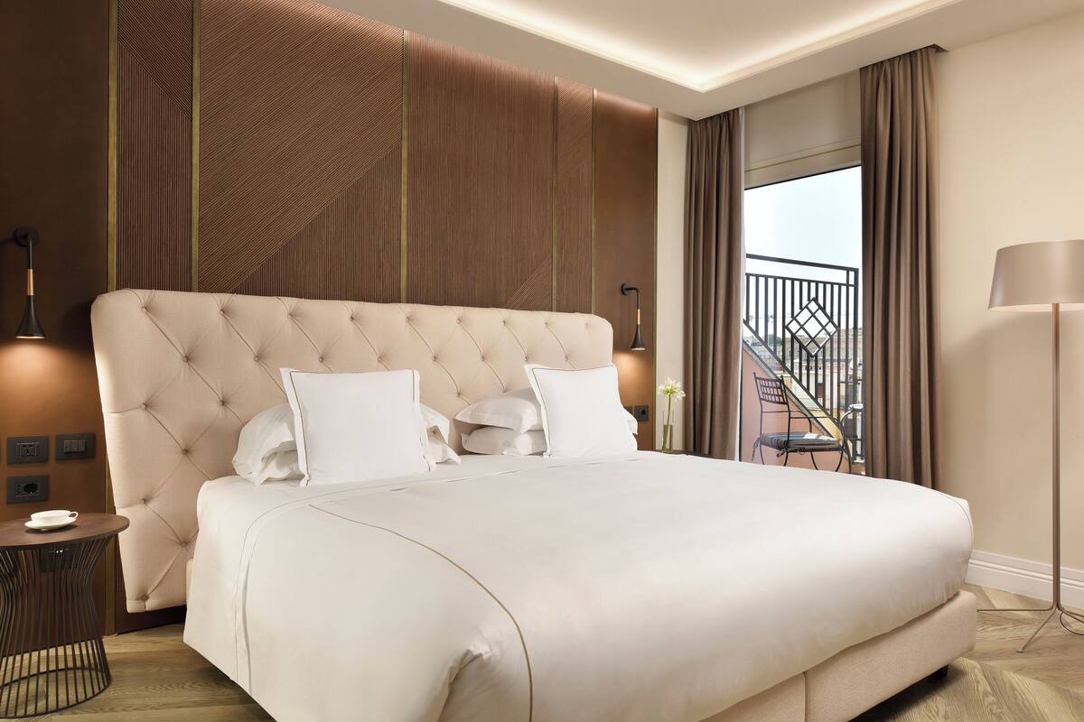 Een groot bed met een gestoffeerde hoofdbord, een nachtkastje met een lamp, en uitzicht op een balkon met een hekwerk.