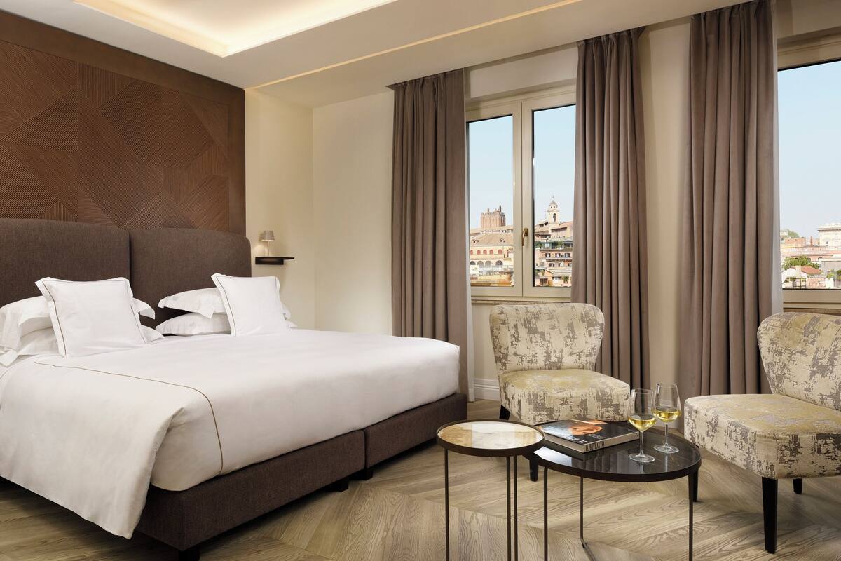 Hotelkamer met een groot bed, twee fauteuils, een tafeltje met glazen en uitzicht op de stad door grote ramen.
