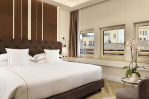 A large bed with white linens, a tufted headboard, and a view of buildings through windows.
