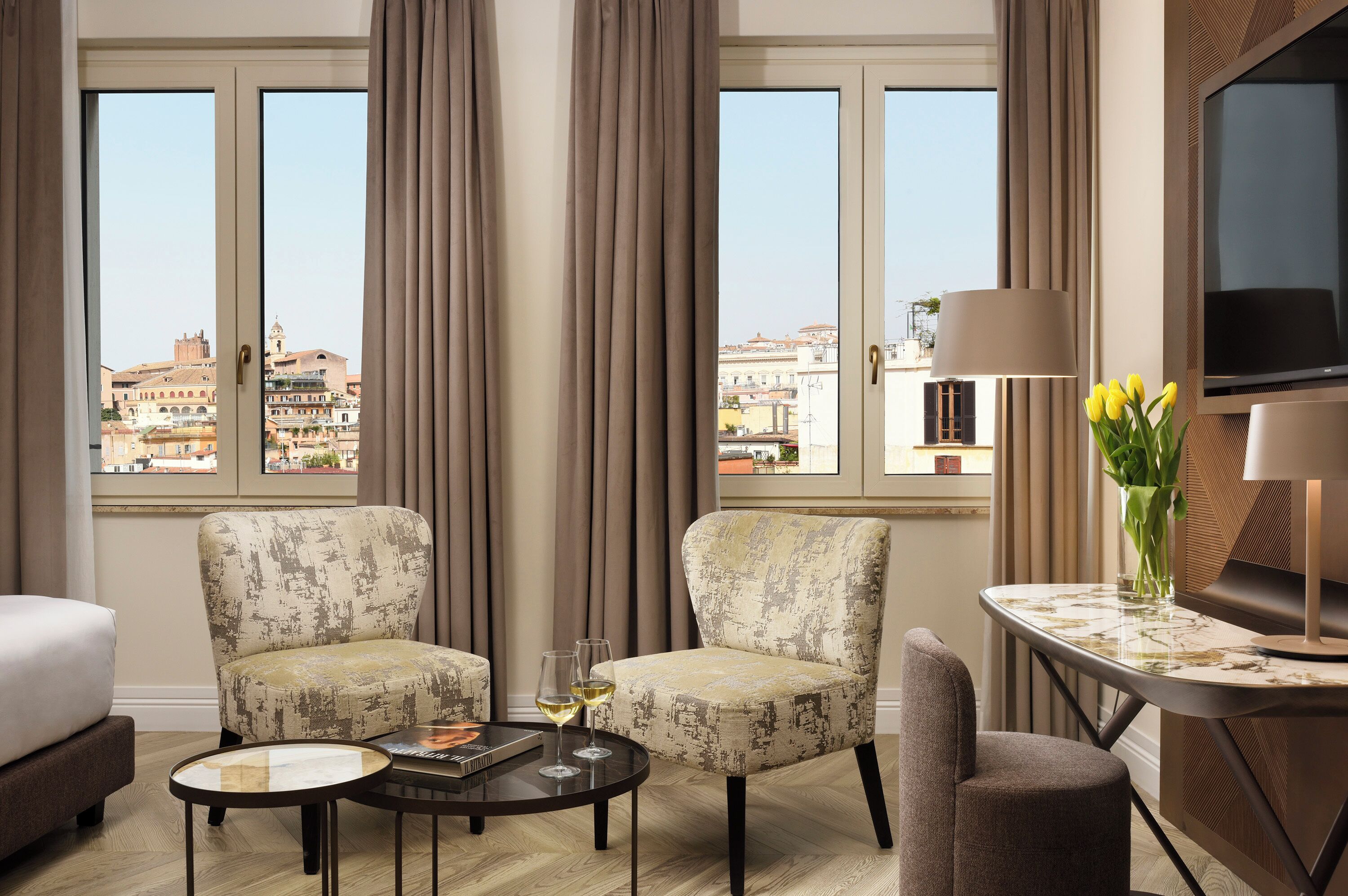 A hotel room with two armchairs, a small round table with a book and glasses, a TV, and a view of the city through large windows.
