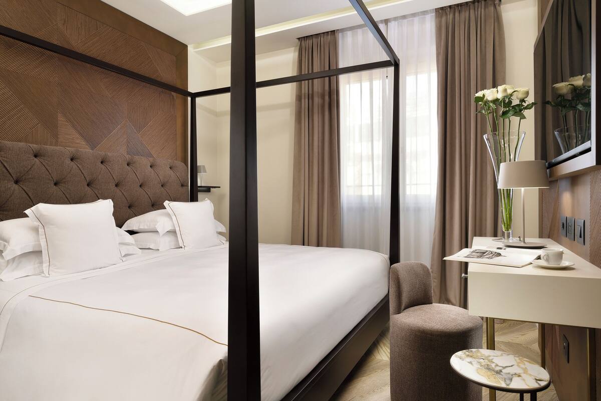 Een hemelbed met witte lakens, een nachtkastje met een lamp, en een klein rond tafeltje met een kop en schoteltje.
