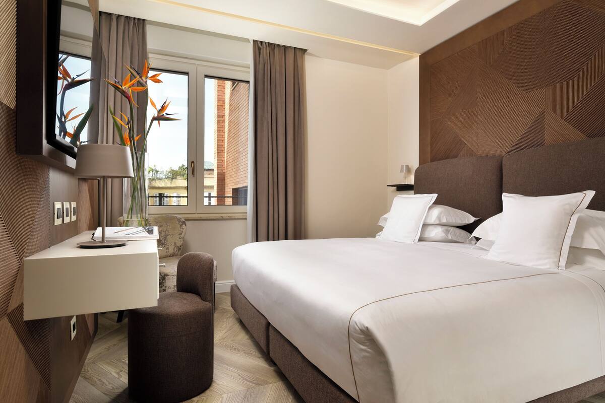 Een moderne hotelkamer met een groot bed, een bureau met lamp, een klein voetenbankje en uitzicht buiten door een raam.
