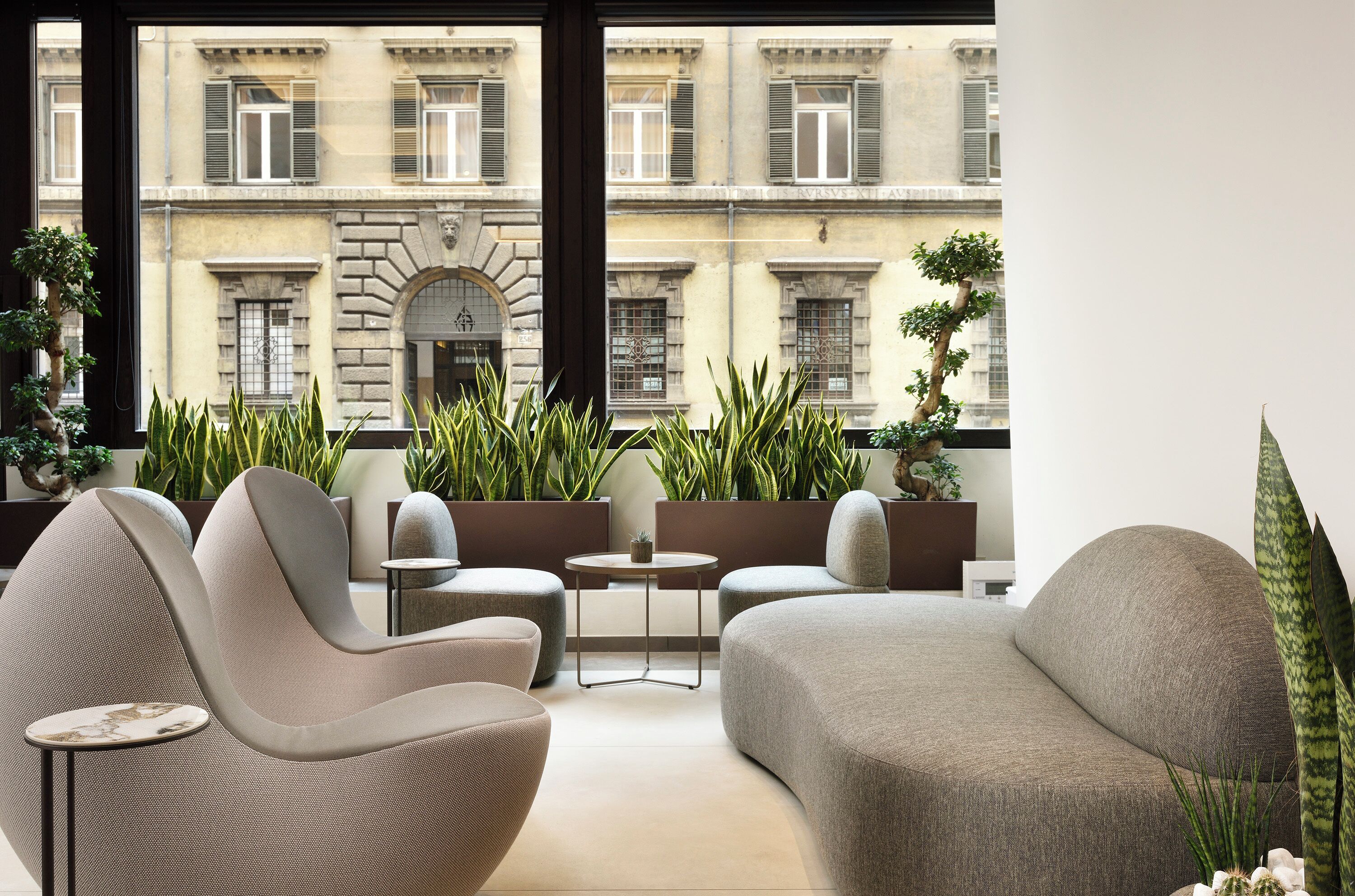 Un salon moderne avec des canapés et une petite table, de grandes fenêtres offrant une vue sur des bâtiments historiques et des plantes en pot.