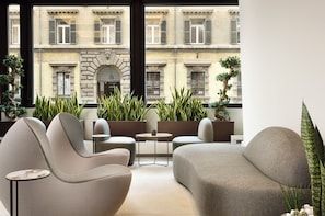 A modern lounge with sofas and a small table, large windows with a view of historic buildings, and potted plants.