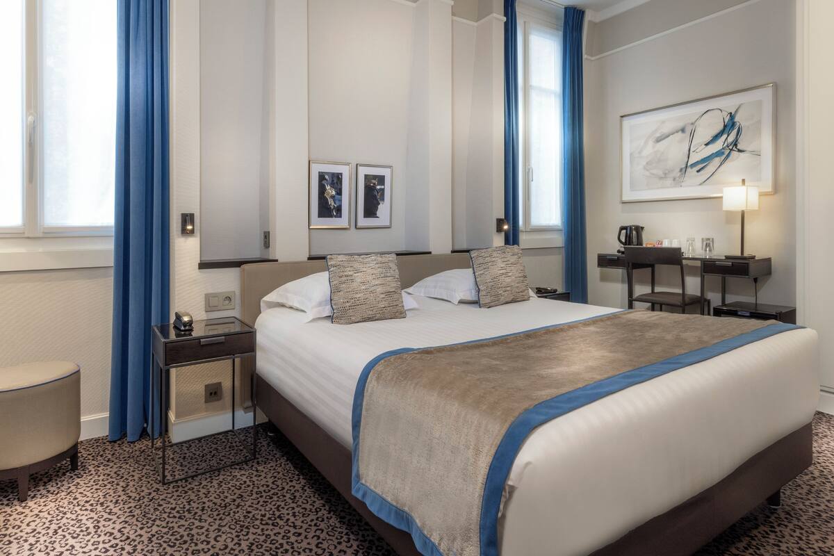 A hotel room with a large bed, a desk, a chair, a nightstand, and a lamp.