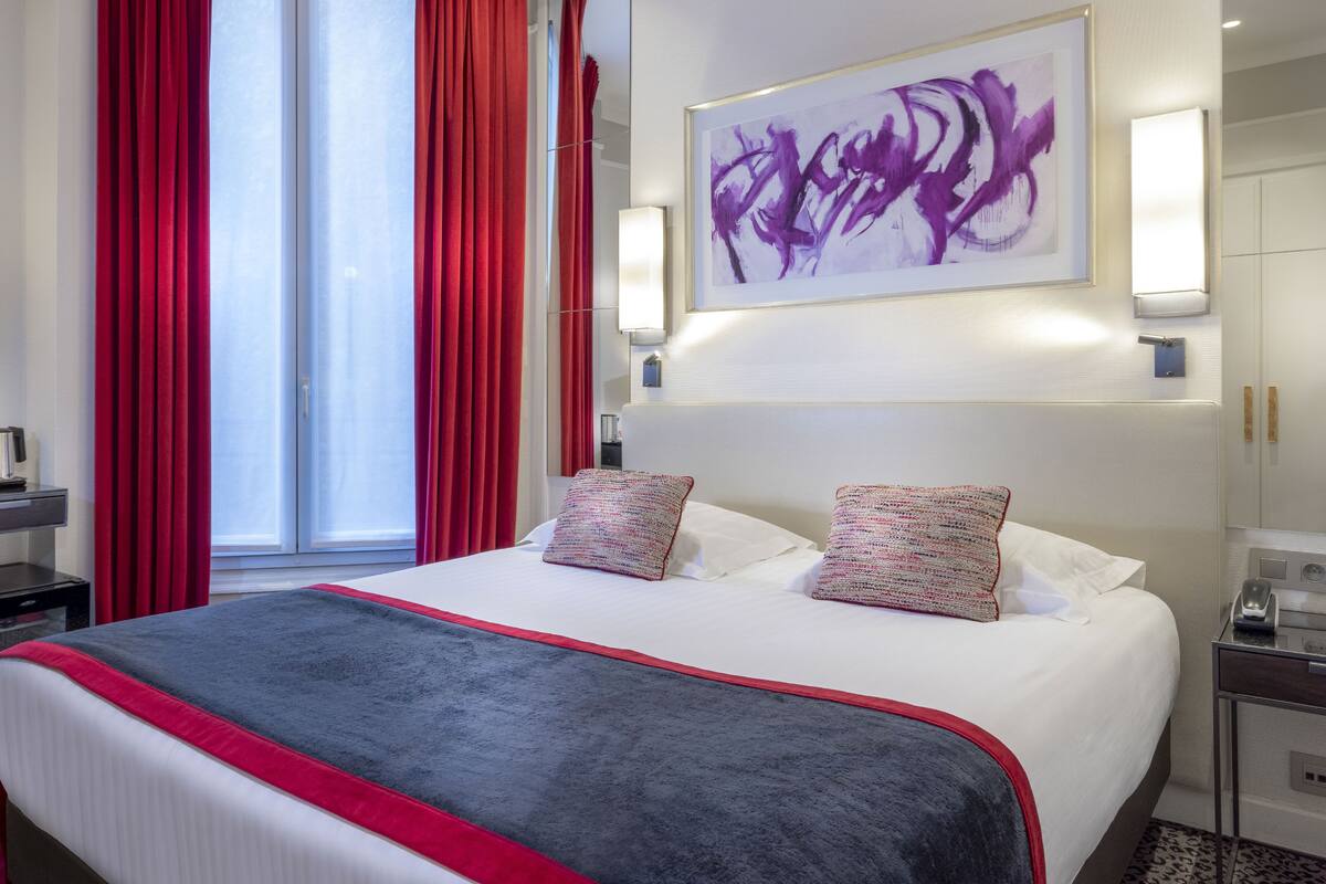 A hotel room with a large bed, red curtains, a wall-mounted abstract painting, and a bedside table with a lamp.