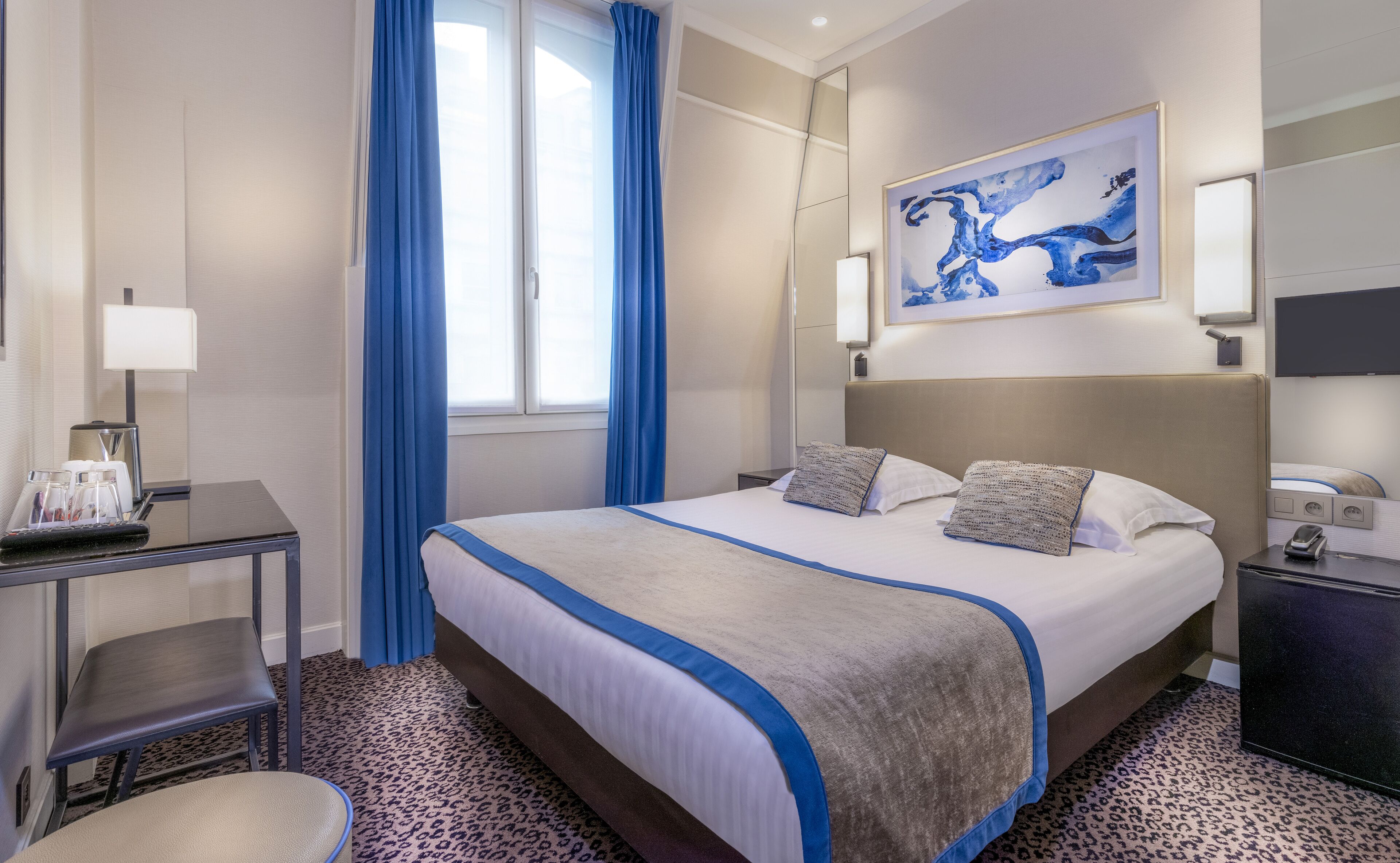A hotel room with a large bed, a desk with a lamp, a chair, and a window with blue curtains.