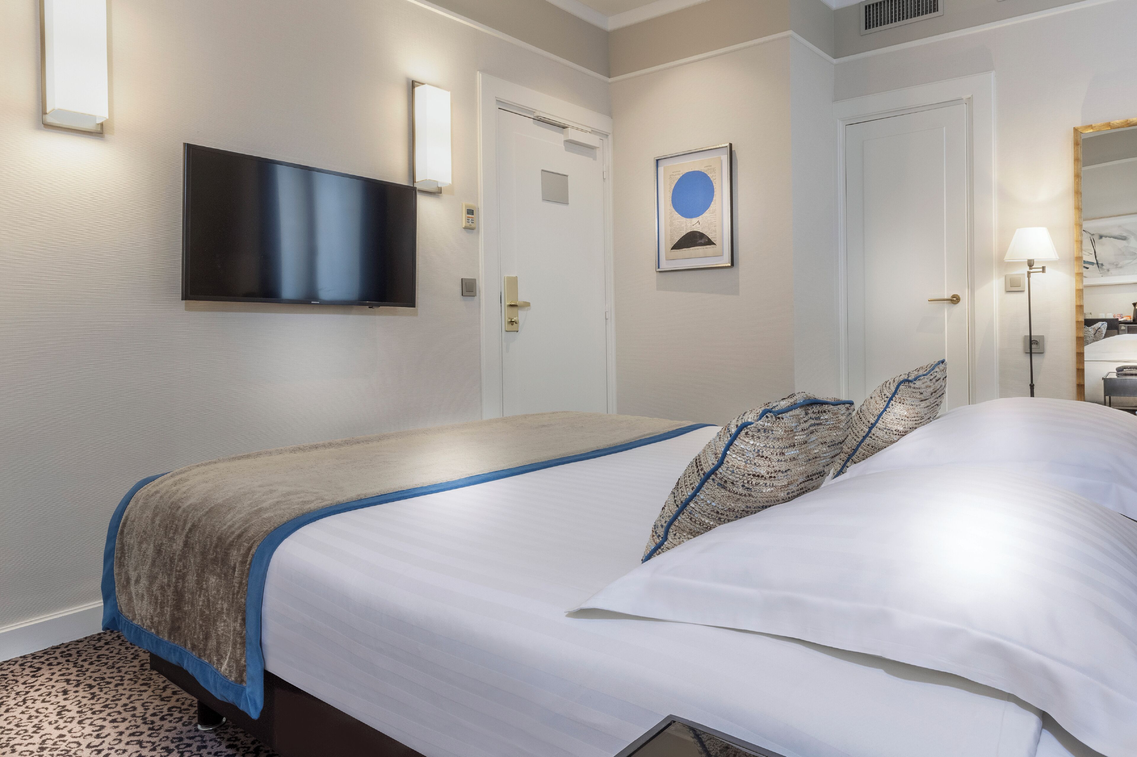 Habitación de hotel con una cama, un televisor de pantalla plana montado en la pared, una puerta con manija y una obra de arte enmarcada en la pared.