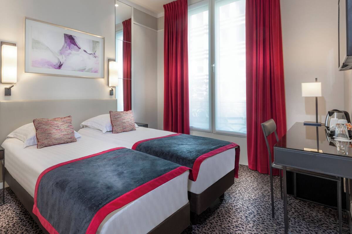 A hotel room with two beds, a desk, a chair, a lamp, and a large window with red curtains.
