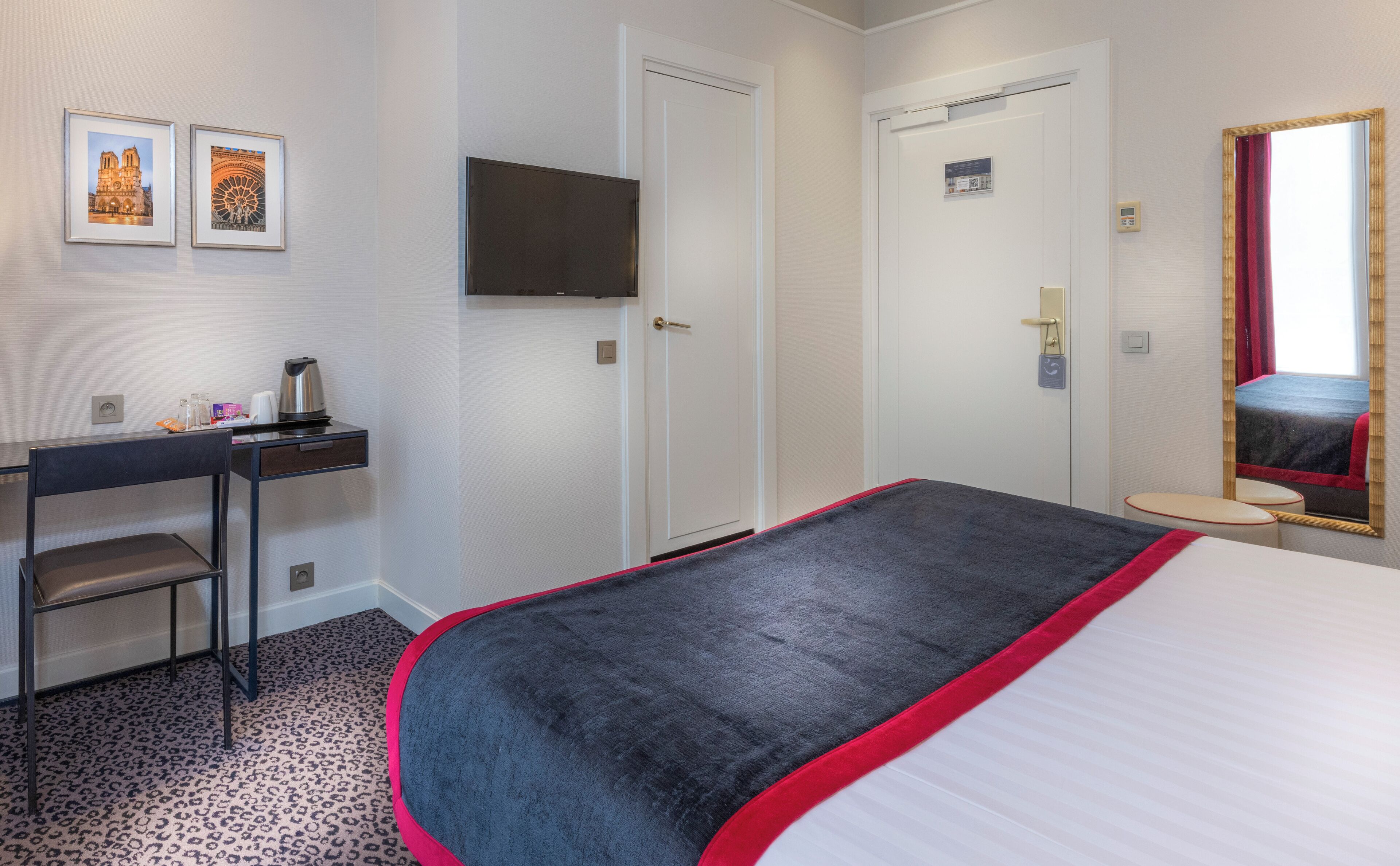 Ein Hotelzimmer mit einem Bett, einem Schreibtisch mit Stuhl, einem Fernseher und zwei gerahmten Bildern an der Wand.