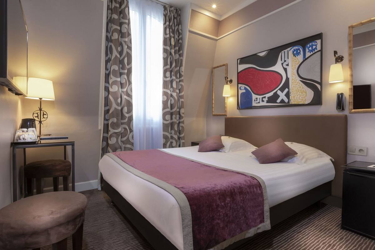 A hotel room with a bed, a desk with a lamp, a chair, a bedside table, and a painting on the wall.