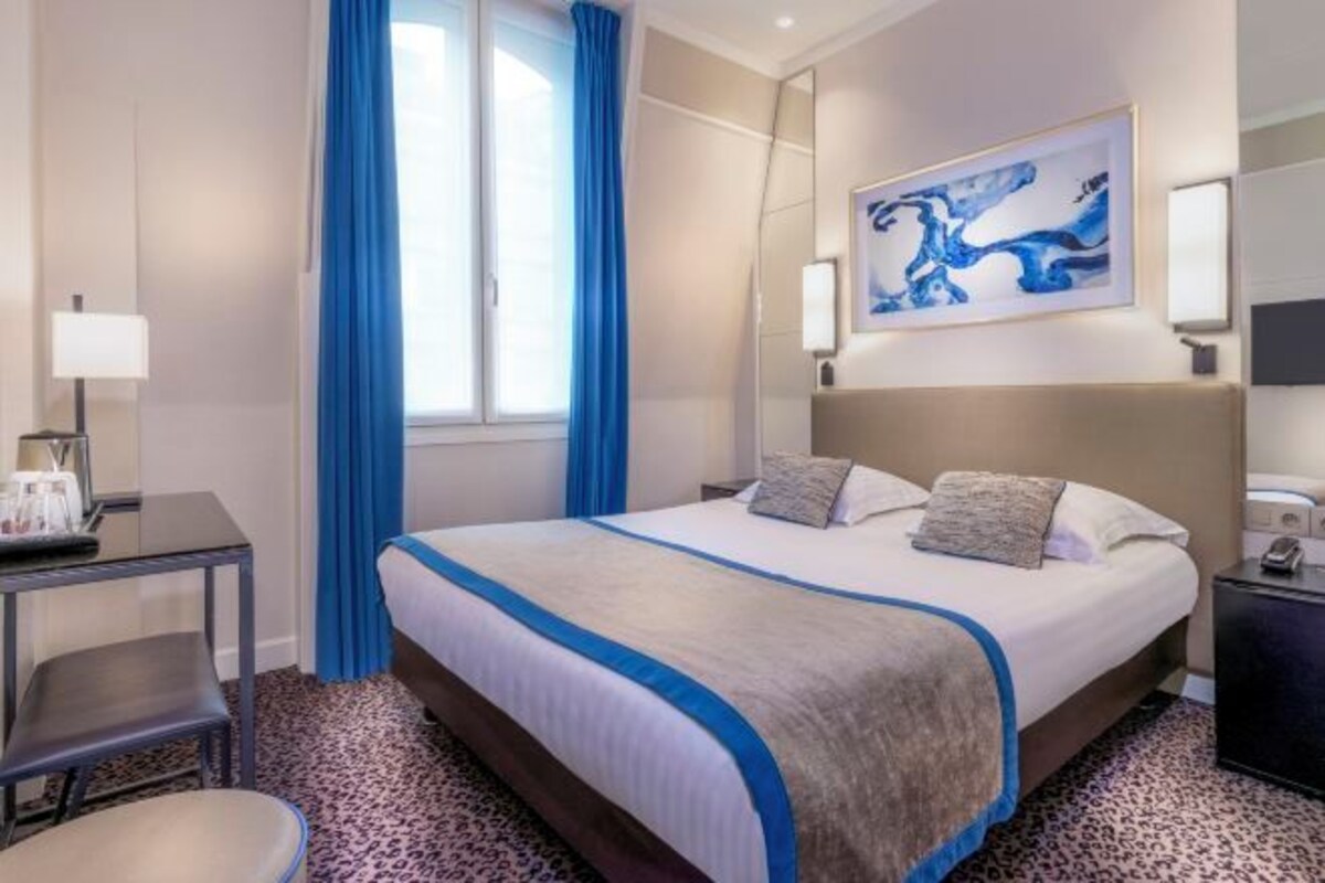 Habitación de hotel con una cama grande, un escritorio con lámpara, una silla y una ventana con cortinas azules.
