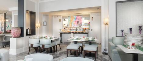 A modern hotel dining area with multiple tables set for breakfast, a bar area, and a seating area with a large painting.