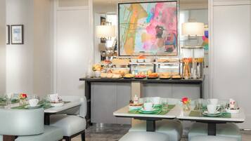 A hotel dining area with tables set for breakfast, featuring a buffet with various food items and a colorful abstract painting above.