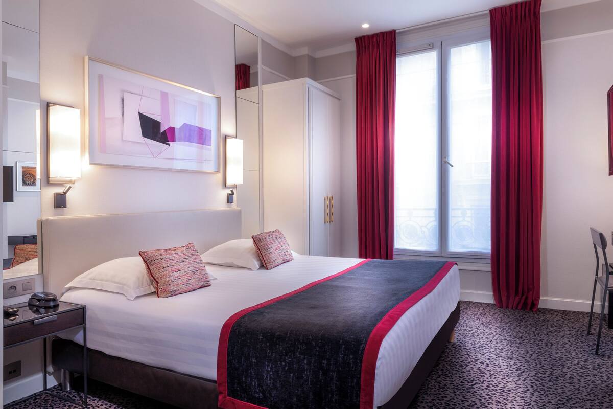 A hotel room with a large bed, a desk, and a chair. There is a window with red curtains and a painting on the wall.