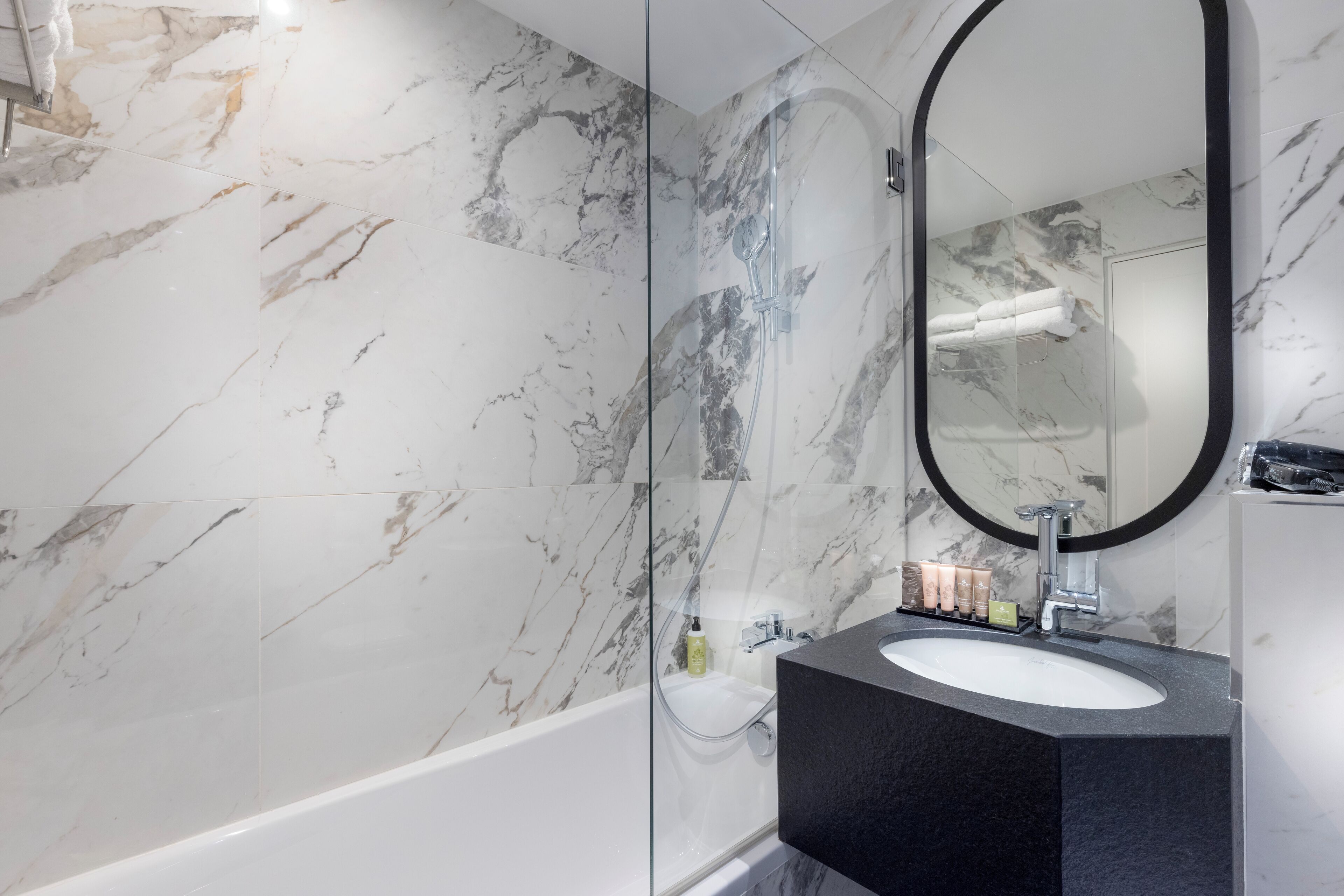 A bathroom with a marble wall, a glass-enclosed shower, a black sink, and a large mirror.