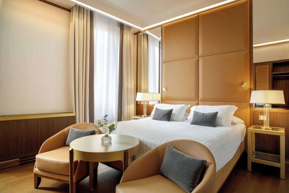 Una camera d'albergo con un letto grande, due comodini con lampade, un tavolino rotondo, due poltrone e uno specchio appeso al muro.