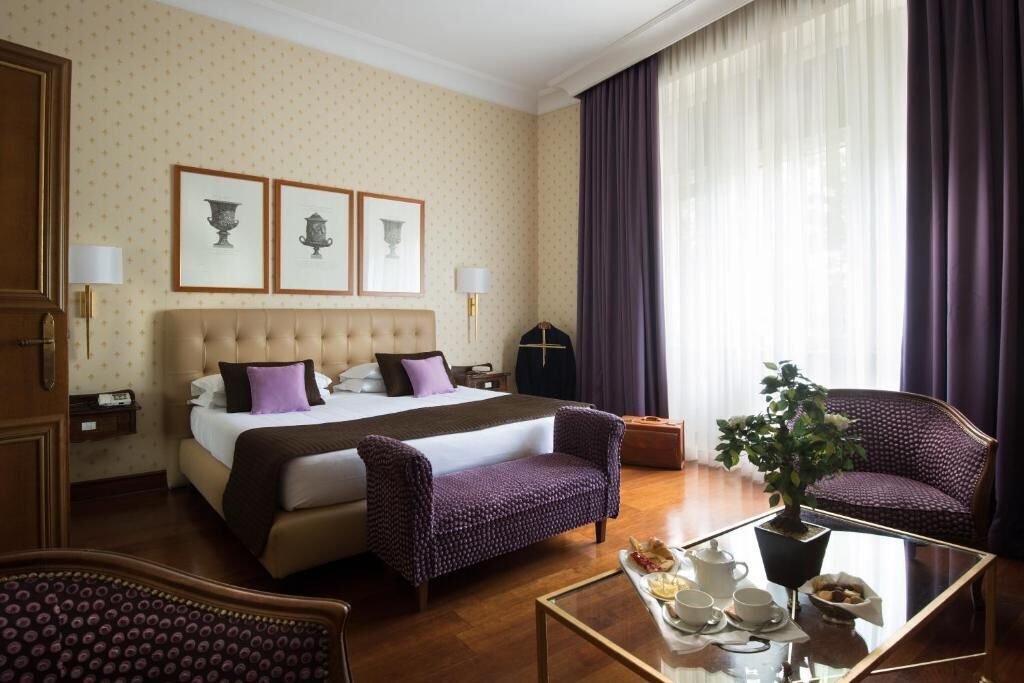 Una habitación de hotel con una cama grande, dos sillones, una mesa de centro de vidrio y una planta en maceta.
