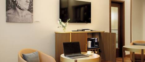 A modern hotel room with a flat-screen TV, a laptop on a small table, a sofa, a coffee machine, and a bookshelf.