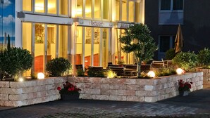 A restaurant with outdoor seating, stone planters with flowers, and a well-lit entrance. - Apart-Hotel Sehnde (Sehnde)