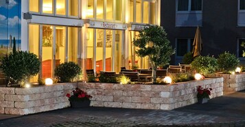A restaurant with outdoor seating, stone planters with flowers, and a well-lit entrance.