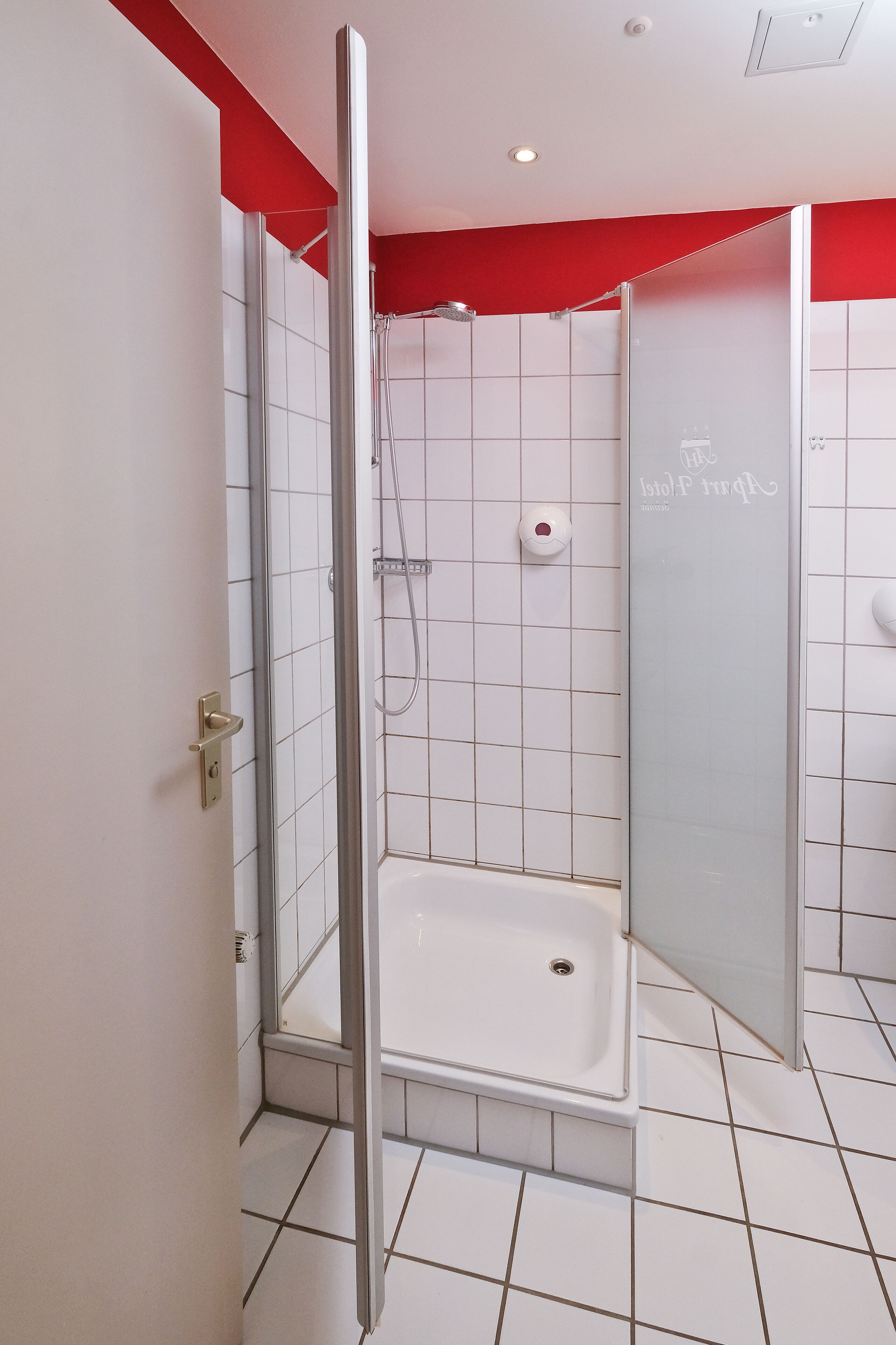 A modern bathroom with a glass shower enclosure, white tiles, and a red accent wall.