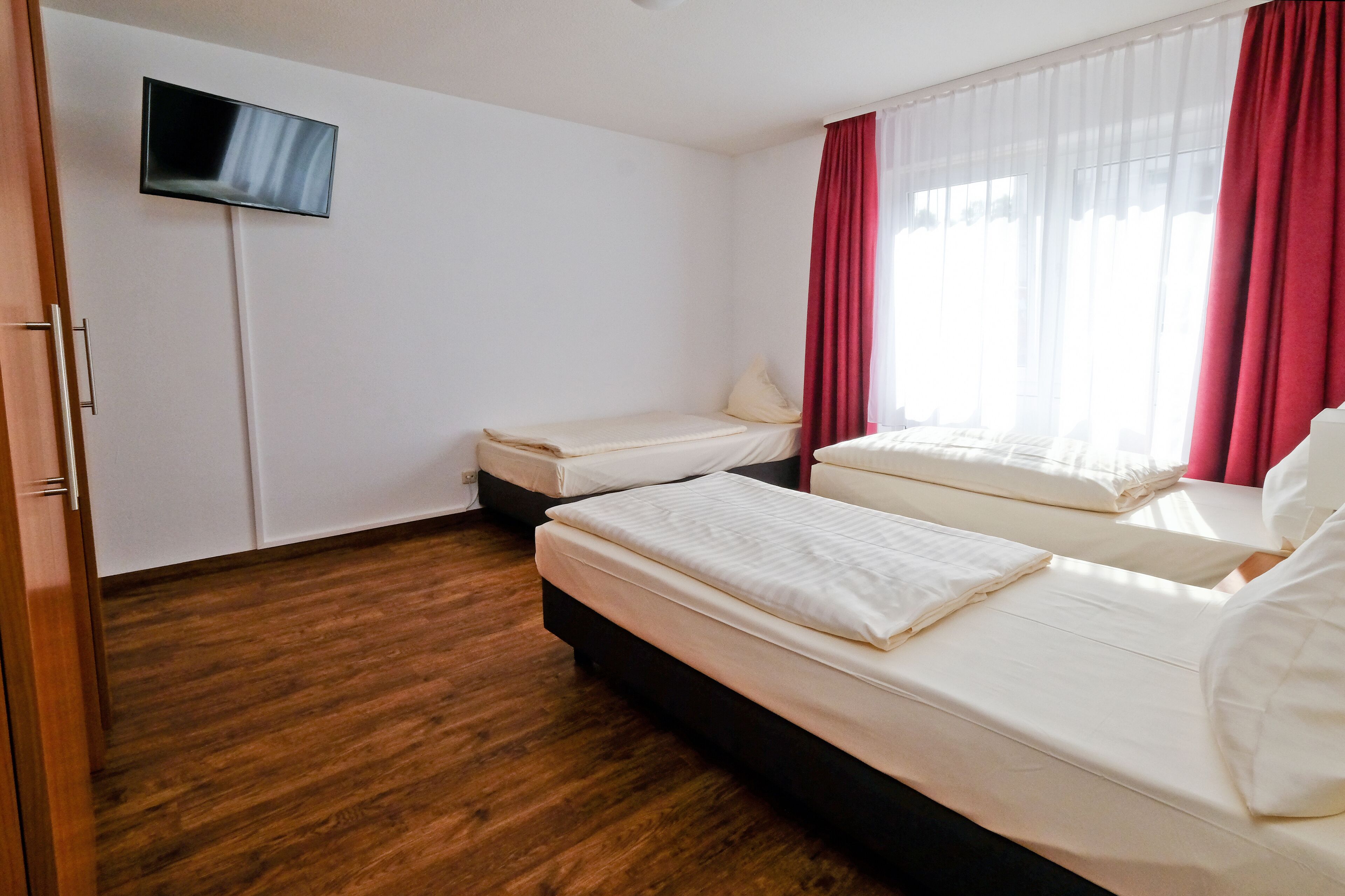 Quarto de hotel com duas camas de solteiro, televisão de tela plana na parede e janela com cortinas vermelhas.