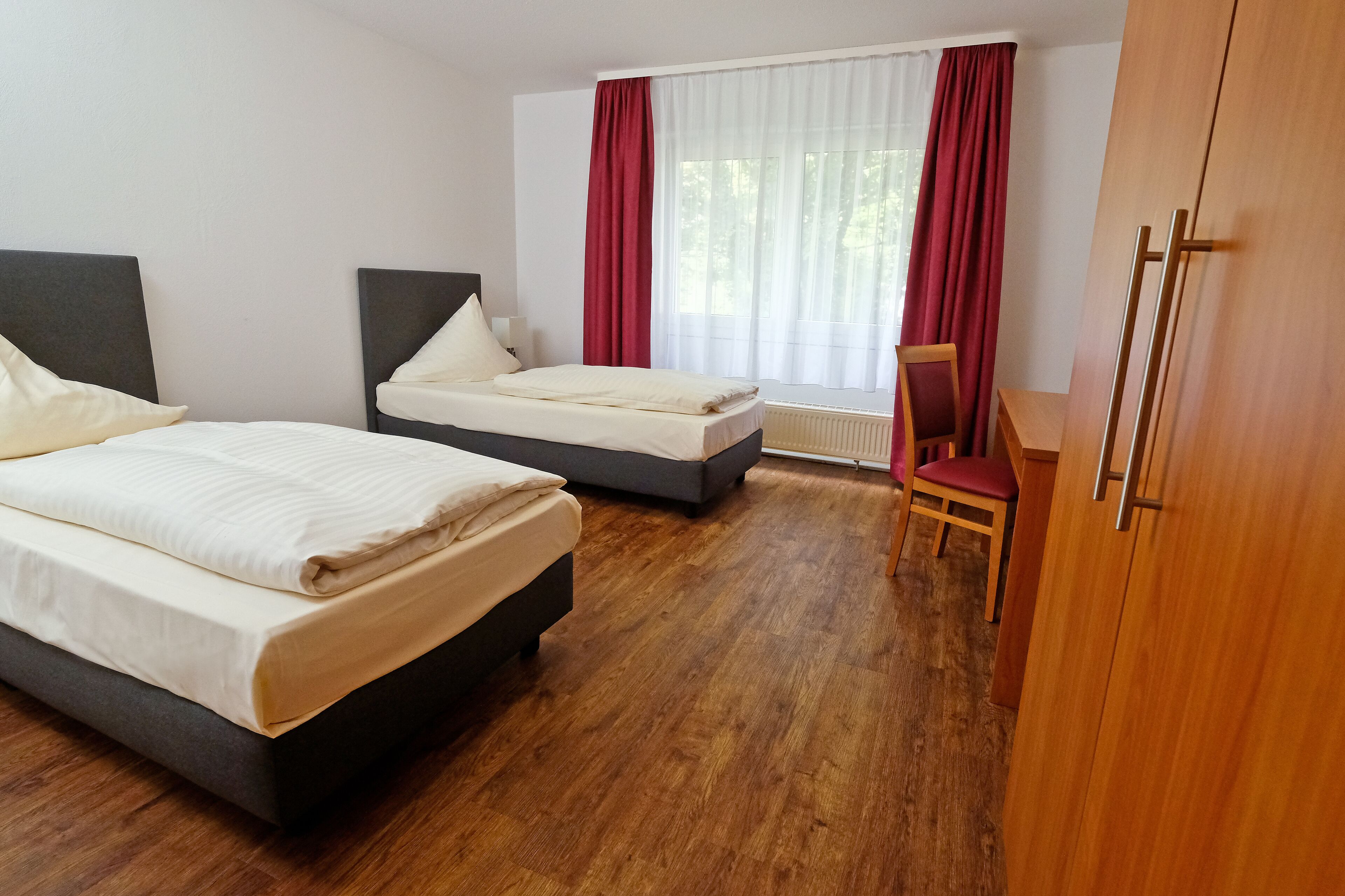 Habitación de hotel con dos camas, un armario de madera, una silla y un ventanal con cortinas.