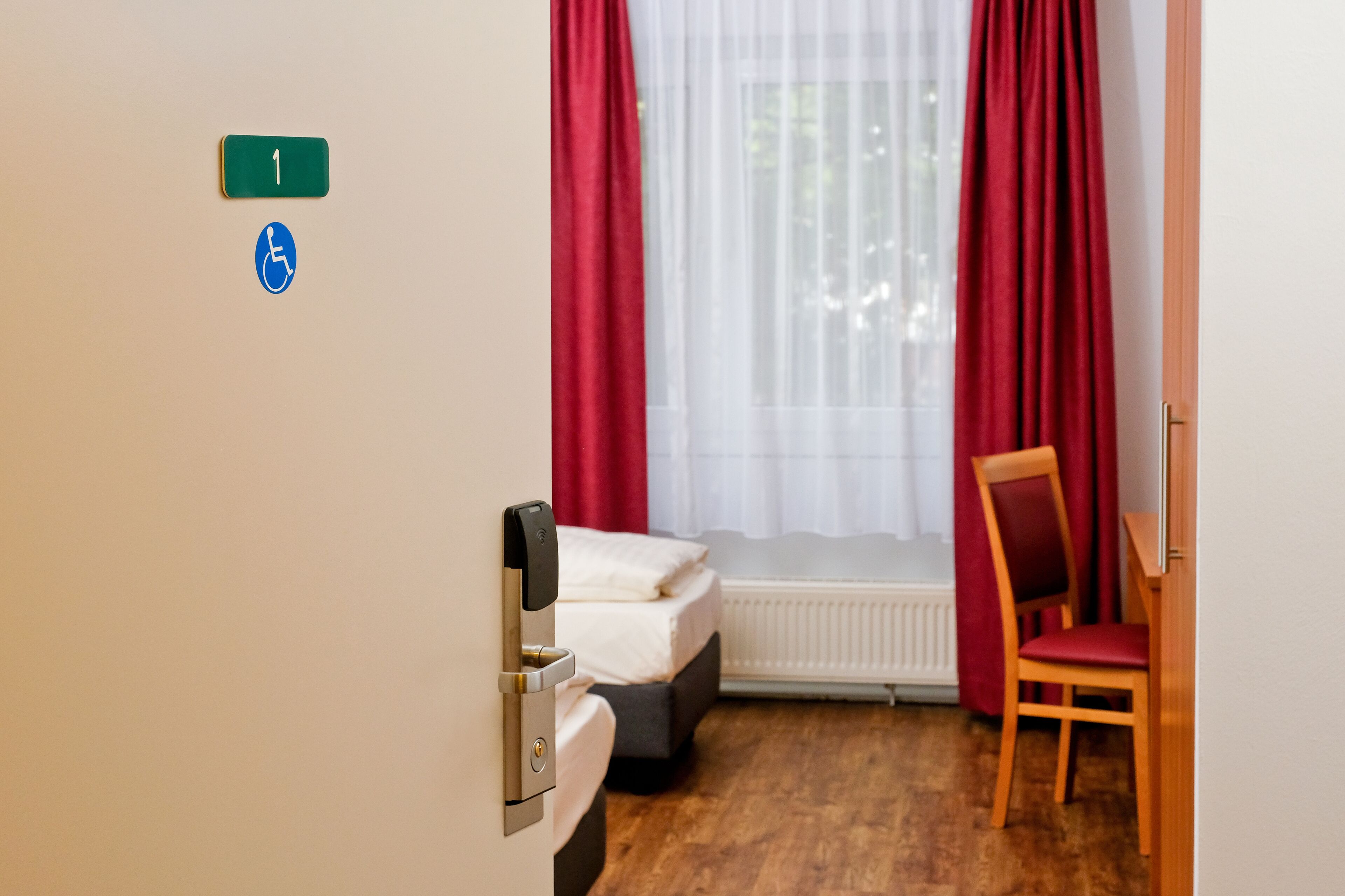A hotel room with a bed, a chair, a window with red curtains, and a sign indicating room number 1 and wheelchair accessibility.
