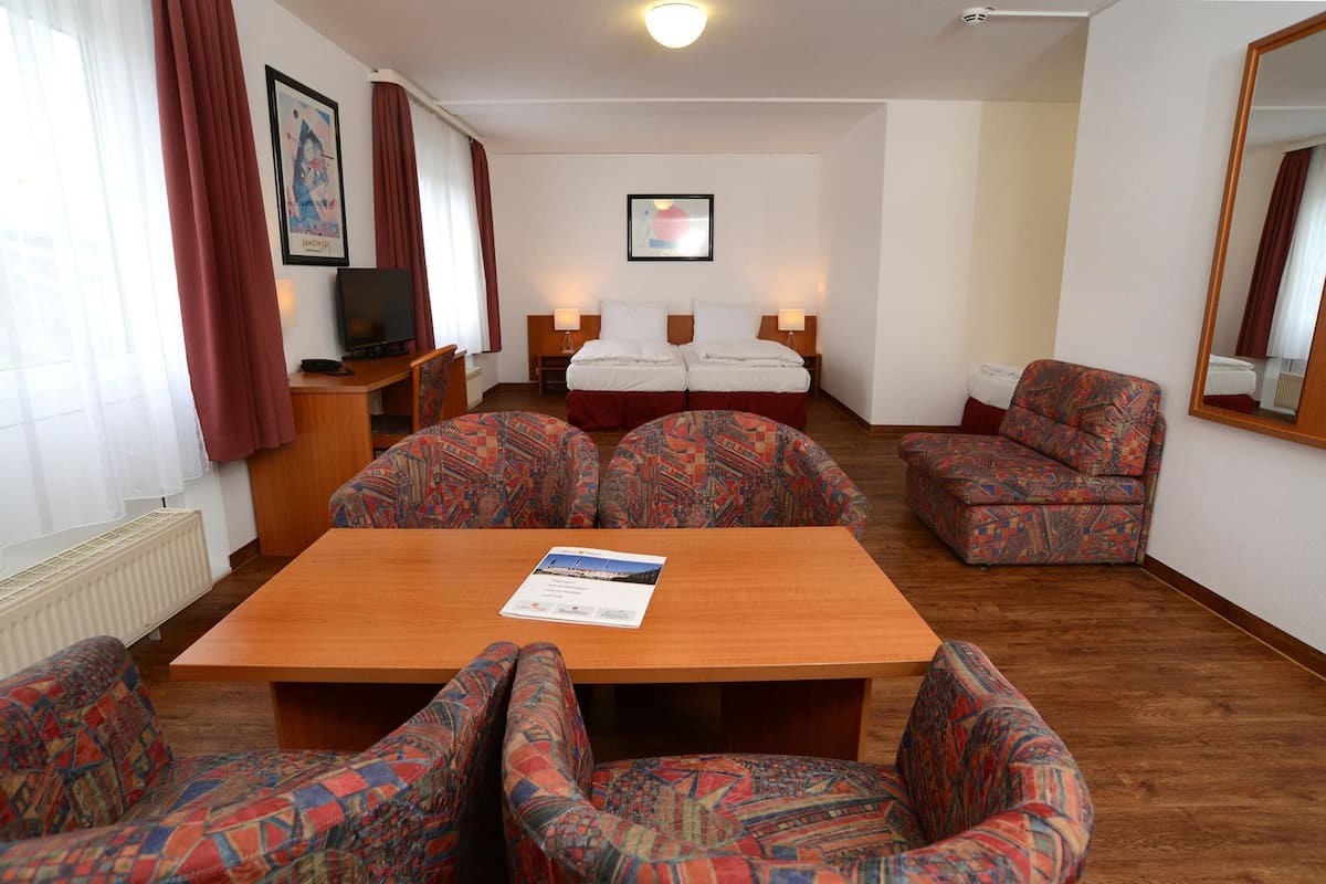 A hotel room with a wooden table, patterned armchairs, a sofa, a bed with two pillows, a TV, and a framed picture on the wall.