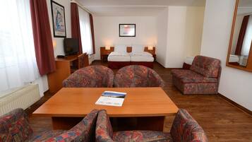 A hotel room with a wooden table, patterned armchairs, a sofa, a bed with two pillows, a TV, and a framed picture on the wall.