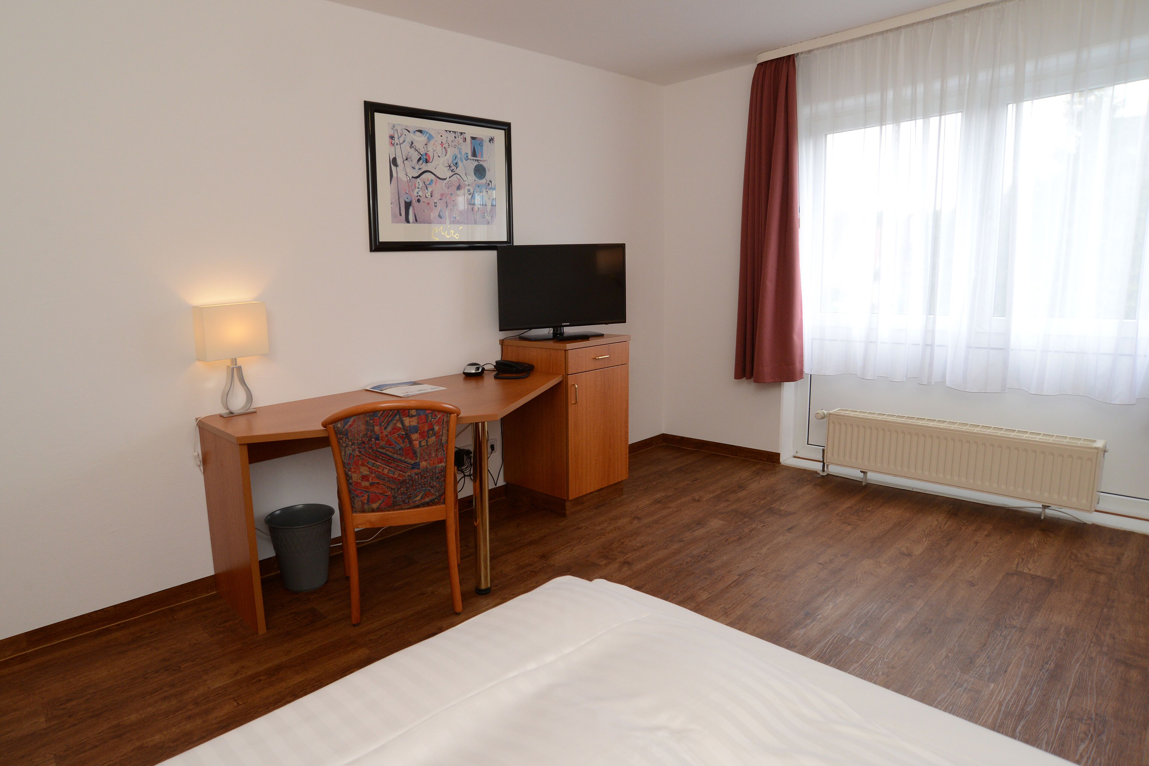 A hotel room with a bed, a desk with a chair, a television, a radiator, and a window with curtains.