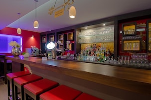 A bar area with red stools, a bar counter, and shelves with glasses and bottles.