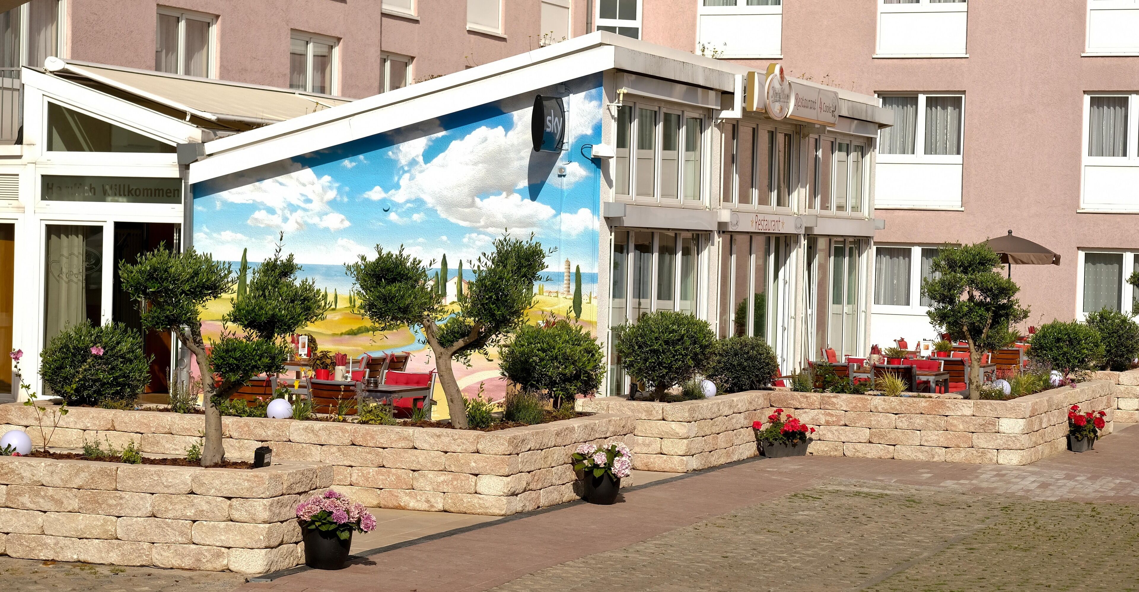 Exterior de hotel com um mural de uma cena de praia, vasos de pedra com flores e área de estar ao ar livre.