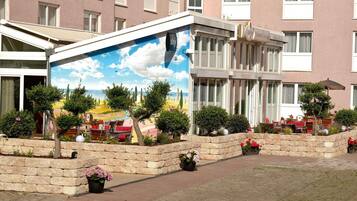 Exterior de hotel com um mural de uma cena de praia, vasos de pedra com flores e área de estar ao ar livre.