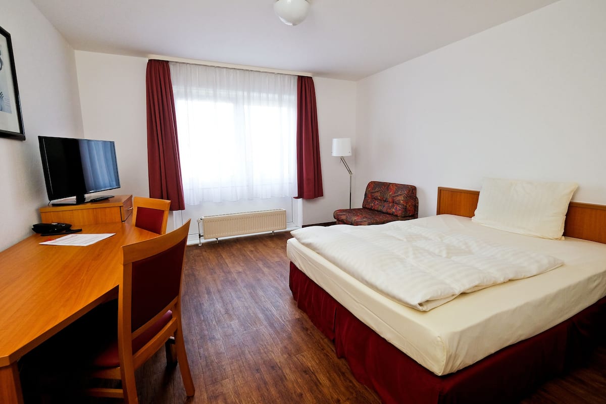 A hotel room with a wooden desk, a television, a bed with red curtains, and a chair.