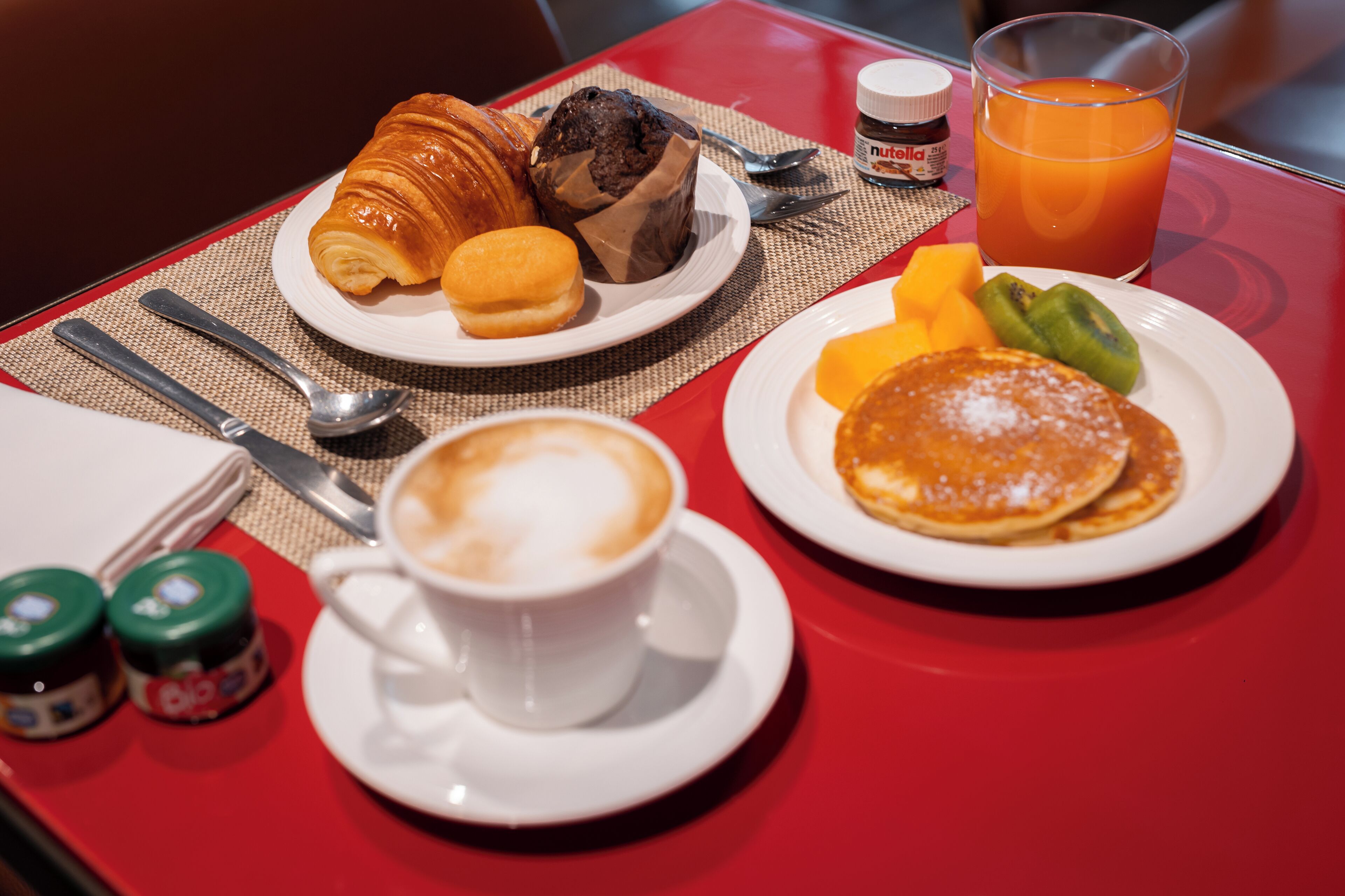 A breakfast setting with croissants, muffins, pancakes, fruit, and a cup of coffee on a red table.