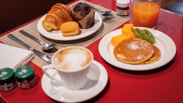 Un desayuno con cruasanes, muffins, tortitas, fruta y una taza de café sobre una mesa roja.