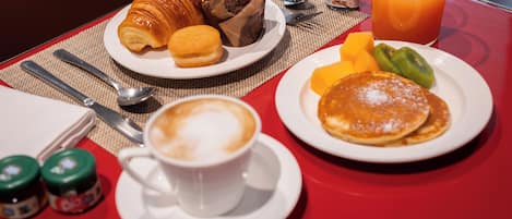 A breakfast setting with croissants, muffins, pancakes, fruit, and a cup of coffee on a red table.