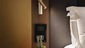 A modern hotel room with a bed, a red side table, a wall-mounted control panel, and a small potted plant.