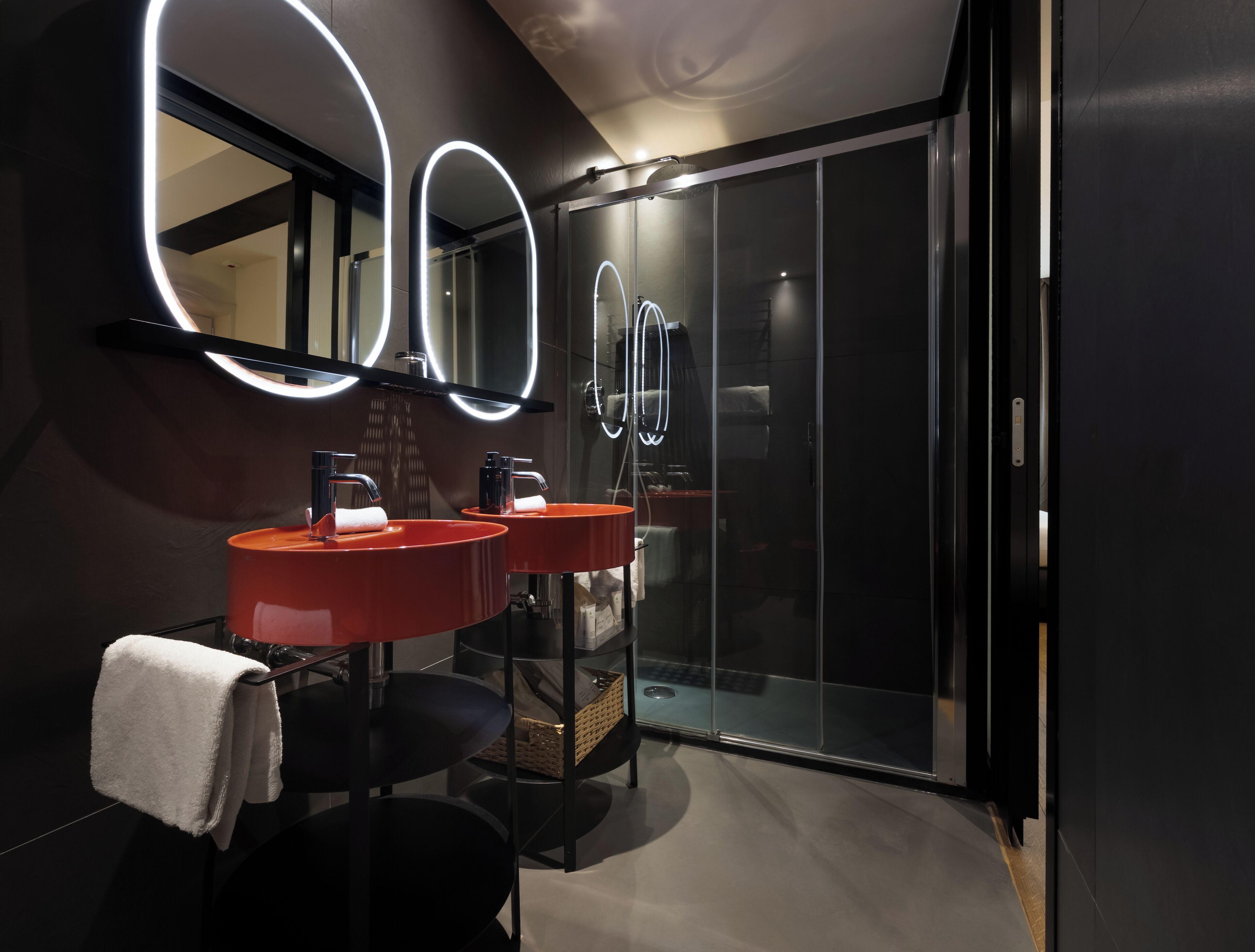Une salle de bain moderne avec deux lavabos rouges, un grand miroir doté de lumières circulaires et une douche en verre.