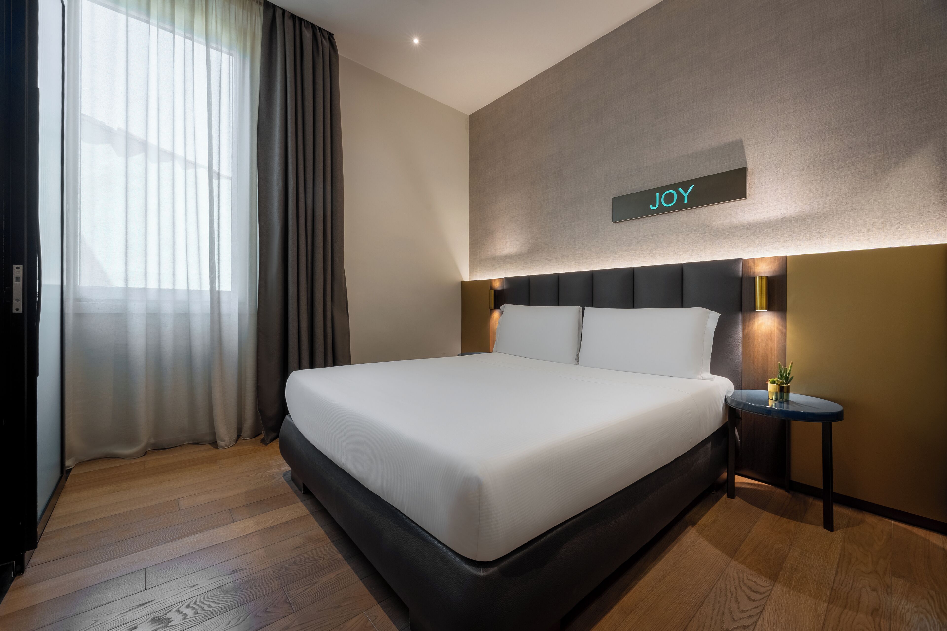 Una habitación de hotel moderna con una cama grande, una mesita de noche con lámpara y un cartel en la pared que dice "JOY".