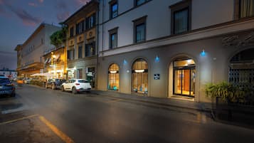 Vista de la calle de un edificio con varias tiendas en la planta baja, incluyendo un mercado. Hay coches estacionados a lo largo de la calle y algunos peatones.