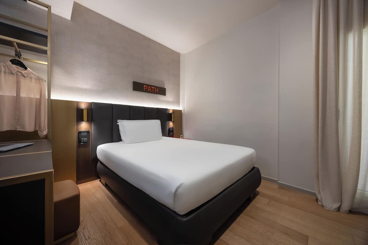 A modern hotel room with a neatly made bed, a bedside table with a lamp, a wardrobe, and a sign above the bed reading "PATH".