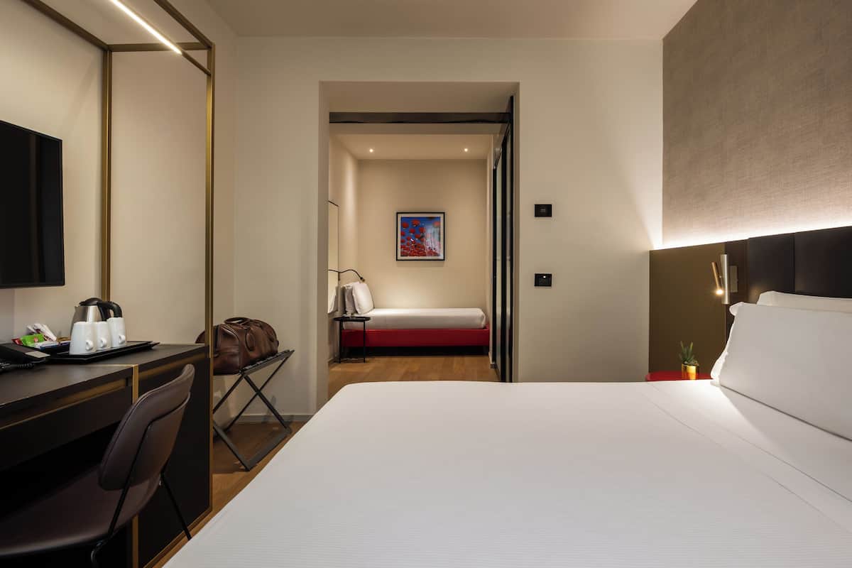 A hotel room with a bed, a desk with a chair, a small table with a plant, and a painting on the wall.
