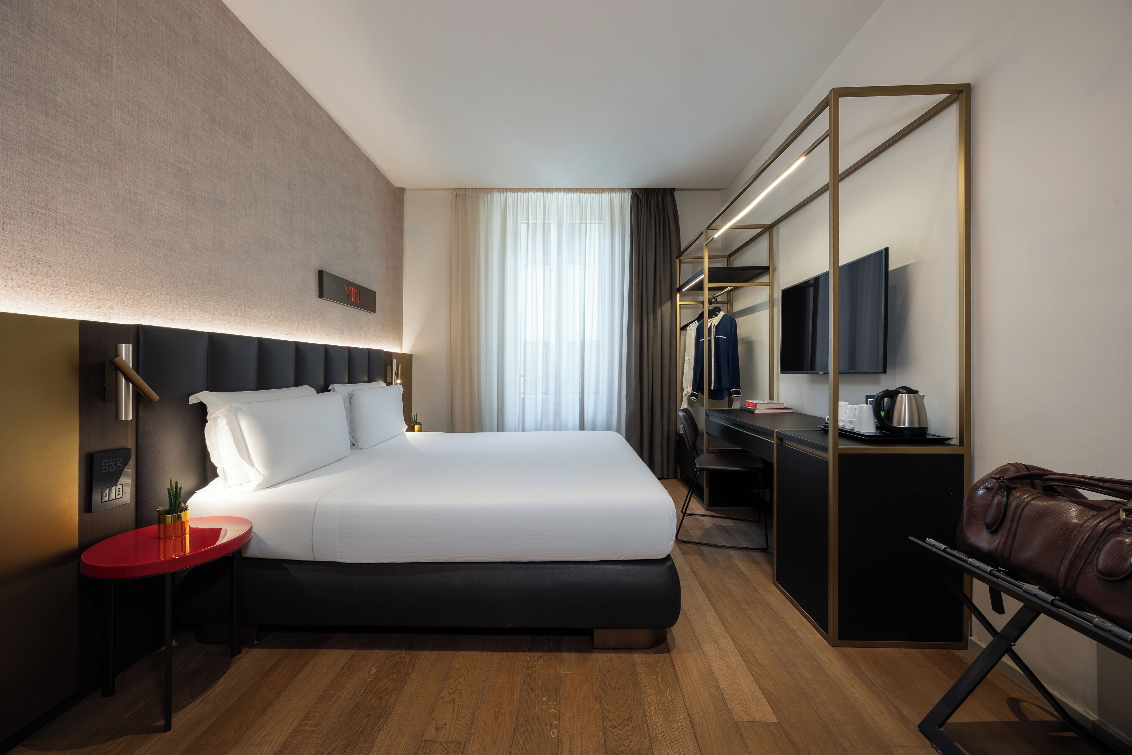 A modern hotel room with a large bed, a desk, a TV, and a leather bag.
