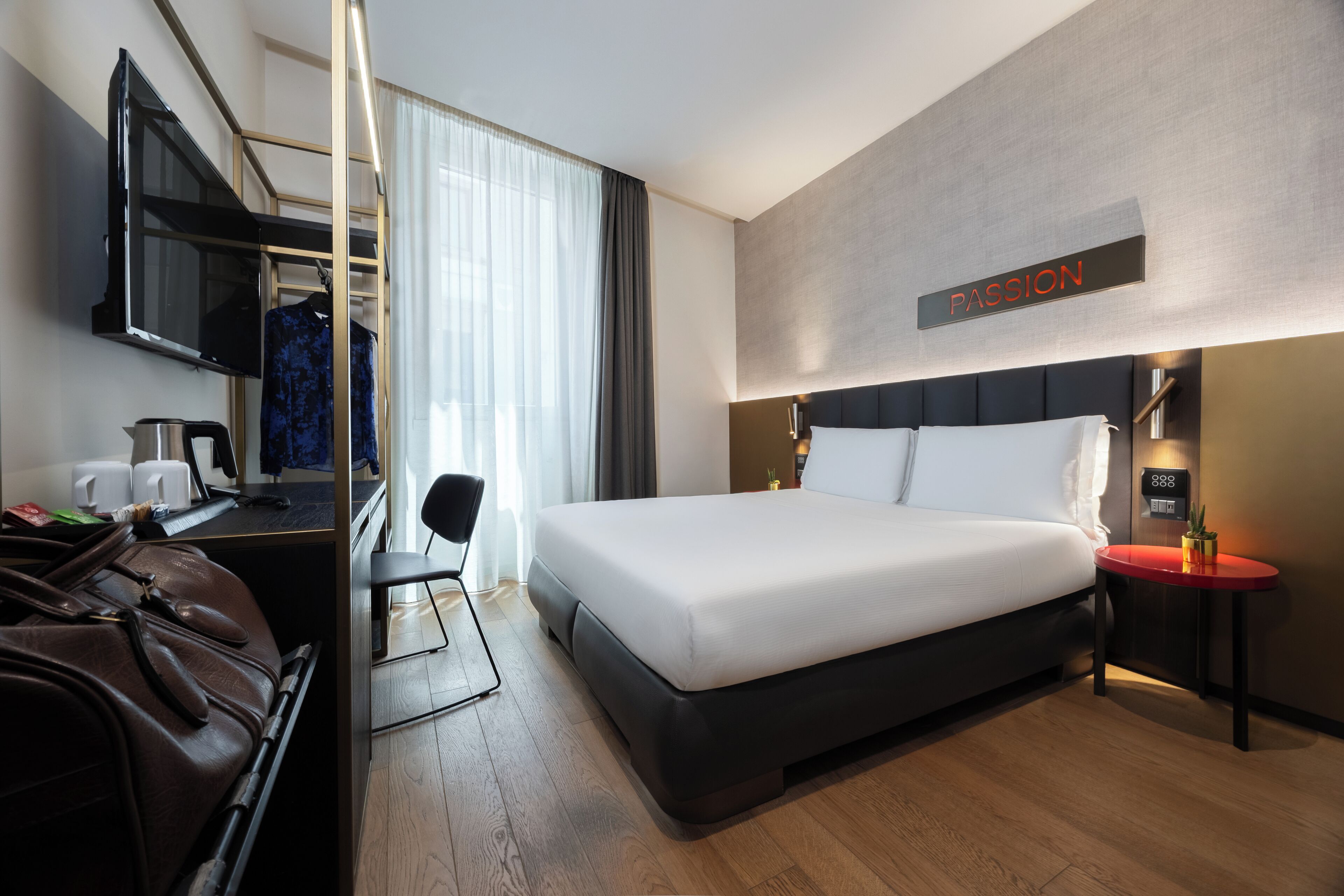 A modern hotel room with a large bed, a desk with a TV, a mirror, and a sign that reads "PASSION".